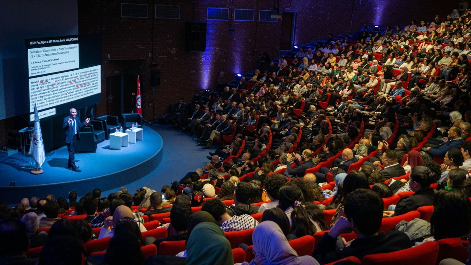 A presenter on a lit stage in front of a full auditorium